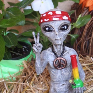 K&ouml;nnte beinhalten: Eine graue Alien-Figur, die ein rot-wei&szlig;es Stirnband und eine Friedenszeichen-Kette tr&auml;gt und einen gr&uuml;nen und gelben Glas-Bong h&auml;lt. Die Figur steht auf einem Strohboden.