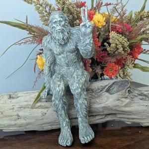 May include: A gray, textured figurine of a hairy, ape-like creature sitting on a piece of driftwood. The creature has a thick coat of fur, a prominent brow, and is holding one hand up in a peace sign gesture with two fingers extended upward.