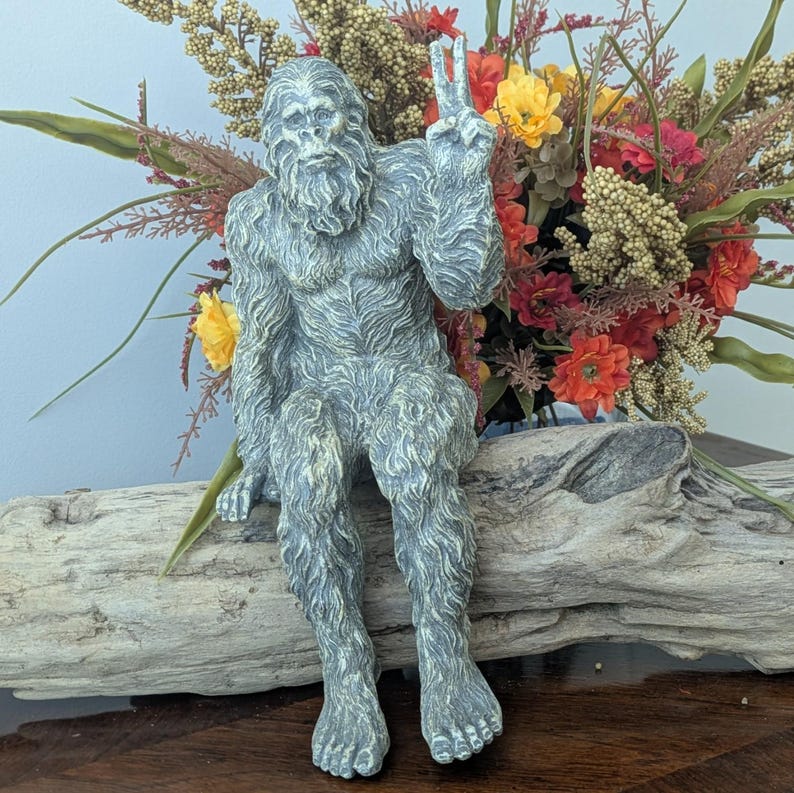 May include: A gray, textured figurine of a hairy, ape-like creature sitting on a piece of driftwood. The creature has a thick coat of fur, a prominent brow, and is holding one hand up in a peace sign gesture with two fingers extended upward.