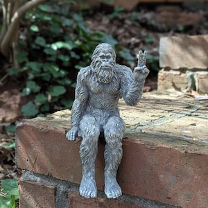 May include: A gray, textured figurine of a seated Bigfoot creature with a shaggy, fur-like appearance. The creature is depicted with a prominent brow, a beard, and its left hand raised in a peace sign gesture, while its legs hang downward.