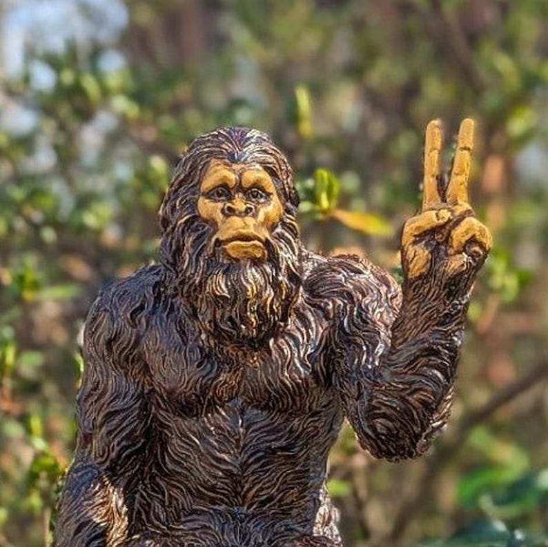 May include: A dark brown, textured figurine of a Sasquatch or Bigfoot creature with a lighter tan face. The creature has thick, shaggy fur and is depicted with its left arm raised, holding up two fingers in a peace sign gesture, facing forward.