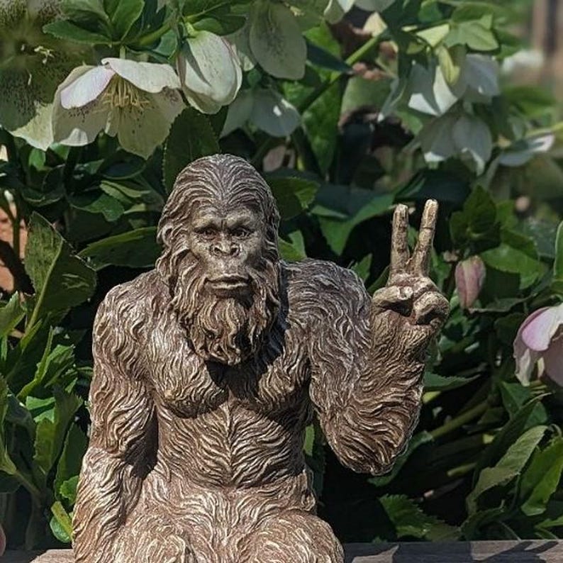 May include: A brown, textured figurine of a hairy, ape-like creature resembling a Bigfoot or Sasquatch. The figure has a detailed, fur-covered body and face, and is depicted with its left hand raised in a peace sign gesture, showing two fingers.