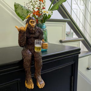 May include: A brown Bigfoot figurine with a beer mug, giving a shaka sign. The figurine has a hairy body, yellow feet and hands, and is sitting on a black surface. A vase of flowers is in the background.