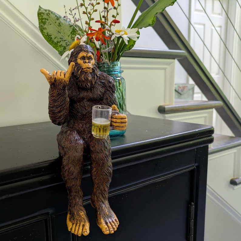 May include: A brown Bigfoot figurine with a beer mug, giving a shaka sign. The figurine has a hairy body, yellow feet and hands, and is sitting on a black surface. A vase of flowers is in the background.