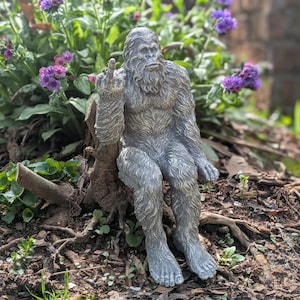 May include: A gray, furry, garden statue of a Bigfoot or Sasquatch sitting on a tree stump with one hand raised in a middle finger gesture.