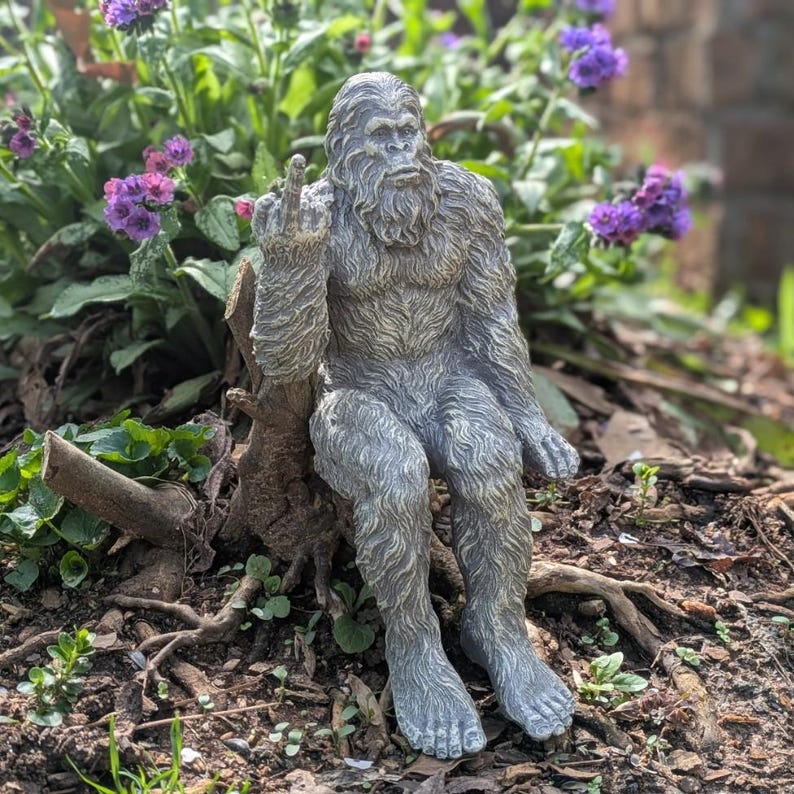 May include: A gray, furry, garden statue of a Bigfoot or Sasquatch sitting on a tree stump with one hand raised in a middle finger gesture.