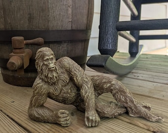 May include: A brown, textured figurine of a hairy, ape-like creature in a reclining pose, resting on one elbow with its legs extended. The figure features detailed fur patterns and a prominent facial structure, crafted in a solid, monochromatic tone.