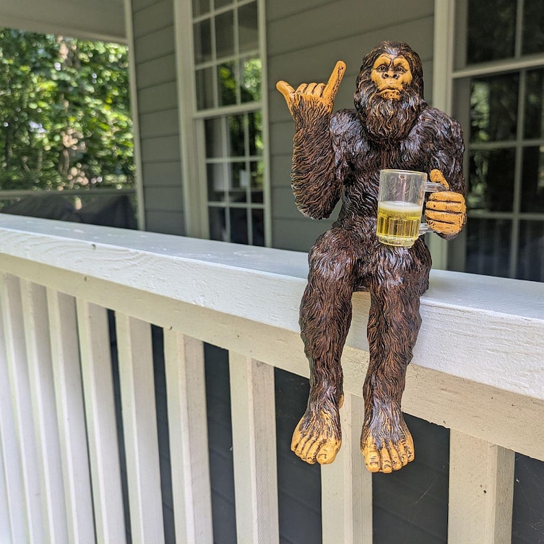 May include: A brown and tan Bigfoot statue with a beer mug, giving a hang loose hand gesture. The statue is sitting on a white railing, with a porch and windows in the background. The beer mug is filled with a yellow liquid.
