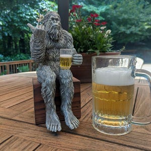May include: A gray Bigfoot statue seated on a wooden block, holding a small glass of beer and giving the middle finger. A large glass beer mug filled with beer sits to the right. The scene is set outdoors on a wooden table.