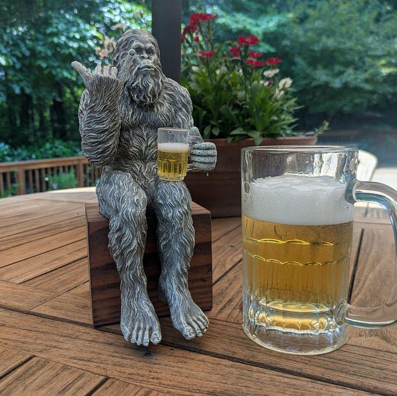 May include: A gray Bigfoot statue seated on a wooden block, holding a small glass of beer and giving the middle finger. A large glass beer mug filled with beer sits to the right. The scene is set outdoors on a wooden table.