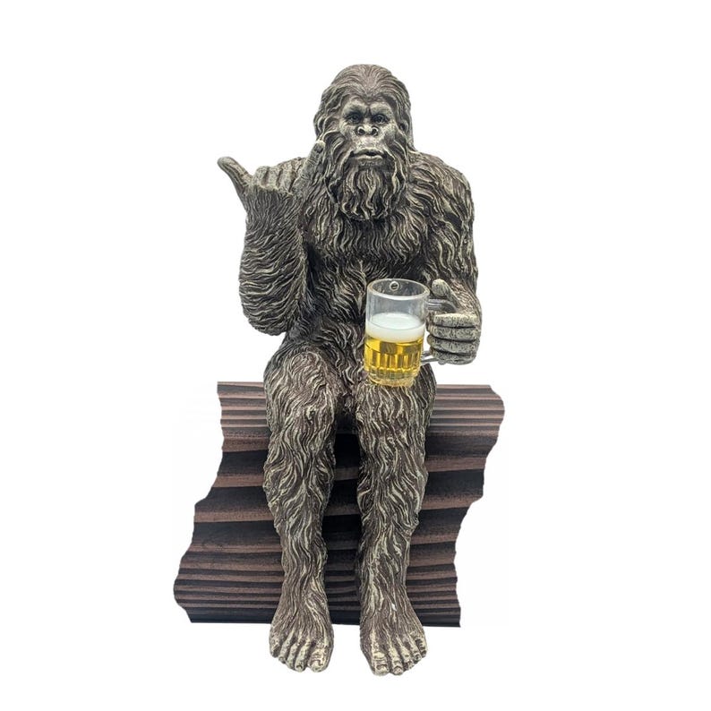 May include: A Bigfoot statue with a beer mug, seated on a wooden bench. The statue is brown and hairy, with a friendly gesture. The beer mug is filled with a golden liquid. A fun, novelty item for home decor.