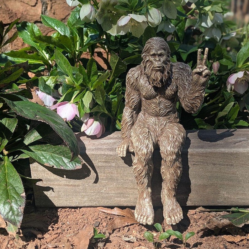 May include: A brown, textured figurine of a hairy, ape-like creature sitting on a ledge. The figure has a thick coat of fur, a prominent brow, and a beard. Its left arm is raised with the index and middle fingers extended in a peace sign gesture.