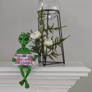 May include: A green alien ornament holding a sign saying "BE MY VALENTINE". The alien is positioned on a white surface, with a glass orb and floral display in the background. The sign has the text "(YOUR NAME HERE)".