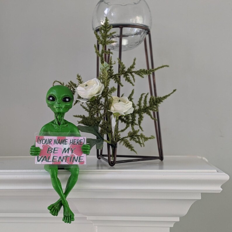 May include: A green alien ornament holding a sign saying "BE MY VALENTINE". The alien is positioned on a white surface, with a glass orb and floral display in the background. The sign has the text "(YOUR NAME HERE)".