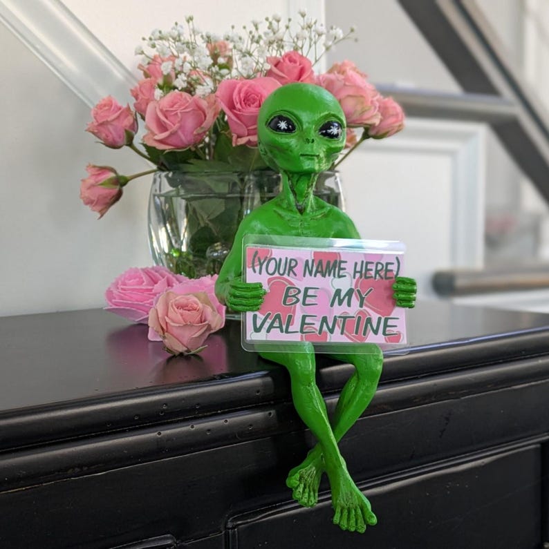 May include: A green alien ornament holding a sign saying "BE MY VALENTINE" with the text "YOUR NAME HERE". The alien is positioned on a black surface, with a vase of pink roses and baby's breath in the background.