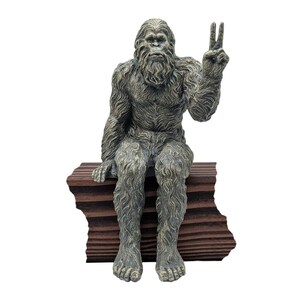 May include: A gray and brown textured figurine of a hairy creature sitting on a dark brown rectangular block. The creature has a thick coat of fur, a prominent face, and its left arm is raised with two fingers extended in a peace sign gesture.