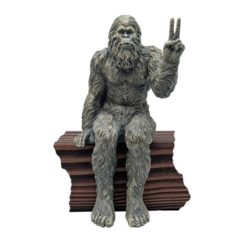 May include: A gray and brown textured figurine of a hairy creature sitting on a dark brown rectangular block. The creature has a thick coat of fur, a prominent face, and its left arm is raised with two fingers extended in a peace sign gesture.