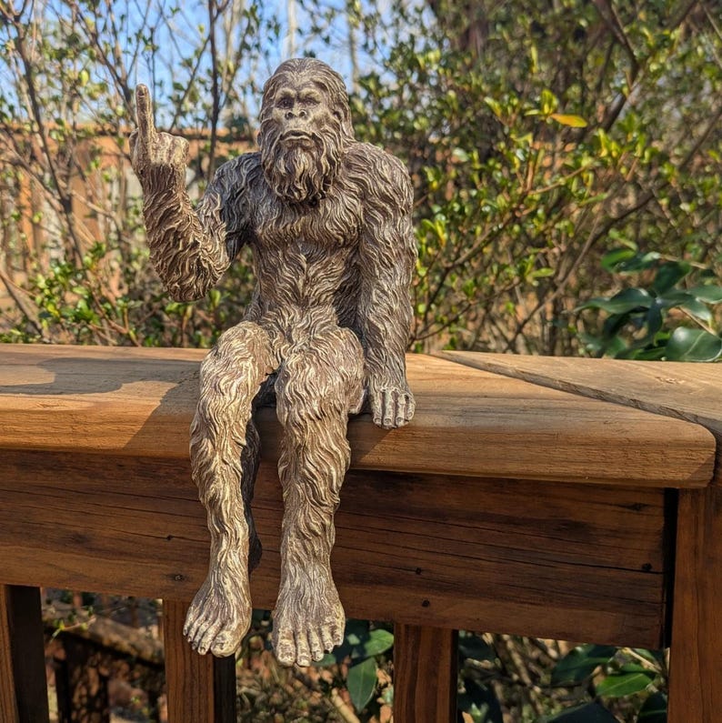 May include: A brown, furry Bigfoot figurine sitting on a wooden railing with one hand raised in a rock and roll hand gesture.