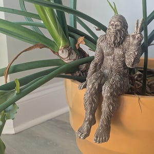 May include: A brown, textured figurine of a hairy, ape-like creature sitting on the edge of a large, solid yellow planter. The creature has a thick beard and is making a peace sign with its left hand, with its legs dangling down the side.