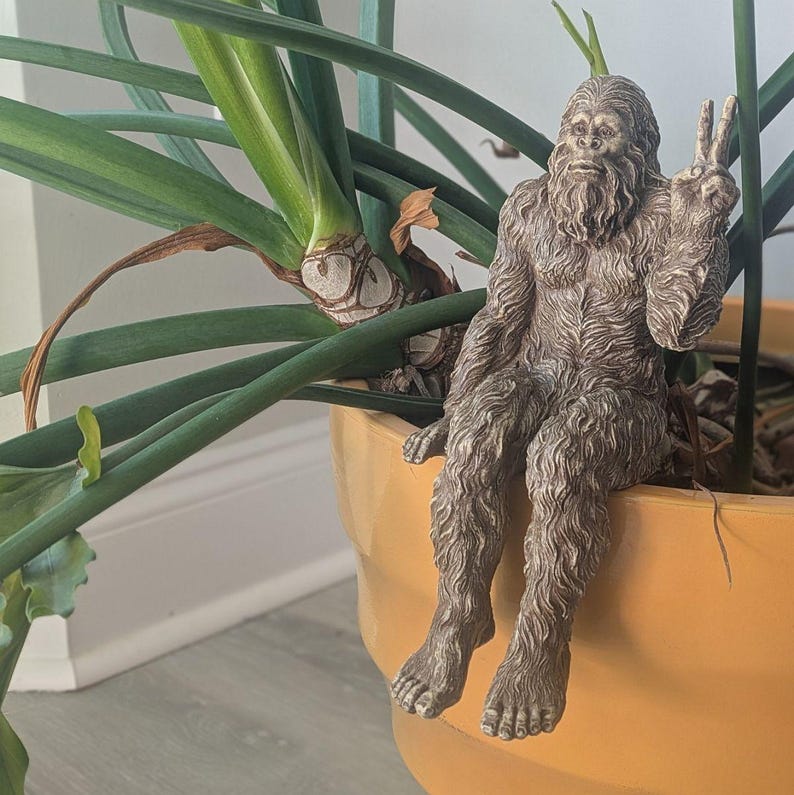 May include: A brown, textured figurine of a hairy, ape-like creature sitting on the edge of a large, solid yellow planter. The creature has a thick beard and is making a peace sign with its left hand, with its legs dangling down the side.