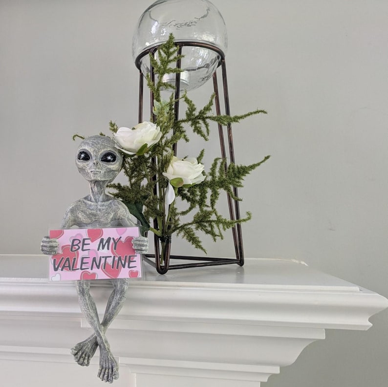 May include: A grey alien ornament holding a pink sign saying "BE MY VALENTINE." The alien is seated on a white surface, legs crossed. Behind it is a decorative metal stand with a glass globe and greenery, a Valentine's Day theme.