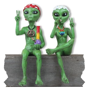 K&ouml;nnte beinhalten: Zwei gr&uuml;ne Alien-Figuren sitzen auf einem Holzsims. Das Alien links tr&auml;gt ein rotes Bandana und h&auml;lt eine Glasbong. Das Alien rechts tr&auml;gt eine wei&szlig;e Blumenkrone und h&auml;lt ein Peace-Zeichen. Beide Aliens haben gro&szlig;e Augen und tragen Peace-Zeichen-Halsketten.
