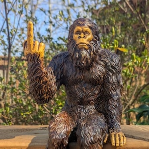 May include: A brown and black figurine of a Bigfoot giving the middle finger. The figurine is sitting on a wooden surface.