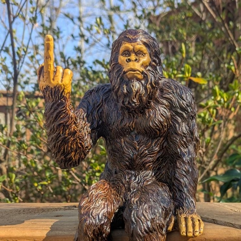 May include: A brown and black figurine of a Bigfoot giving the middle finger. The figurine is sitting on a wooden surface.