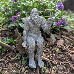May include: A gray, textured statue of a hairy, ape-like creature sitting down. The figure has a thick beard, fur-covered body, and is holding one hand up in a peace sign gesture. The sculpture features detailed, realistic fur patterns throughout.
