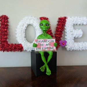 May include: A green alien figure holding a sign saying "Brandon Be My Valentine." The alien is positioned on a black box, with the word "LOVE" in the background, decorated with red and white tinsel.