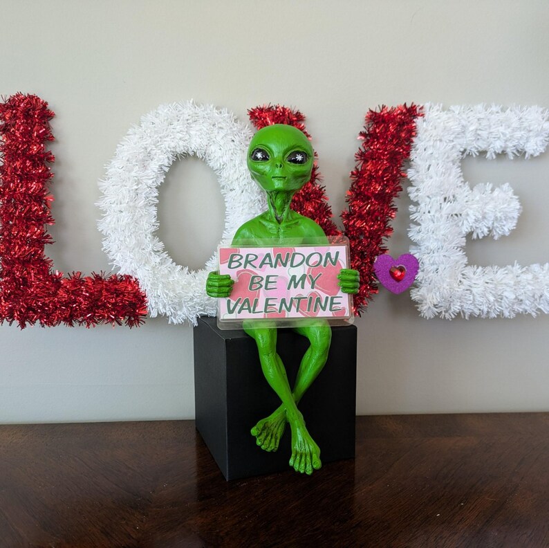 May include: A green alien figure holding a sign saying "Brandon Be My Valentine." The alien is positioned on a black box, with the word "LOVE" in the background, decorated with red and white tinsel.