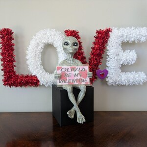May include: A Valentine's Day decoration featuring a grey alien figurine holding a sign that reads "Olivia Be My Valentine." The alien is seated on a black cube, with the word "LOVE" spelled out in red and white tinsel letters in the background.