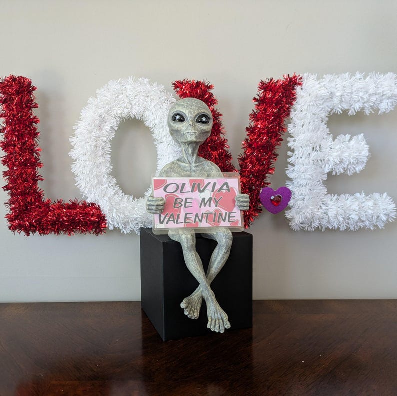 May include: A Valentine's Day decoration featuring a grey alien figurine holding a sign that reads "Olivia Be My Valentine." The alien is seated on a black cube, with the word "LOVE" spelled out in red and white tinsel letters in the background.
