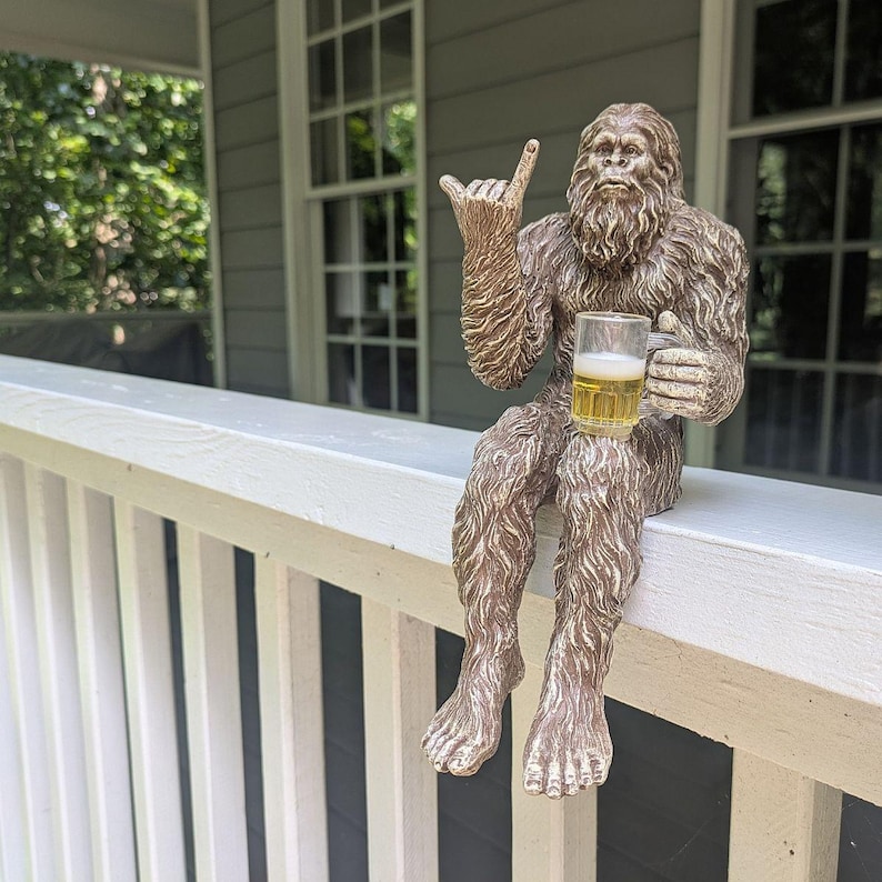 May include: A bronze-colored Bigfoot statue with a full beard and shaggy fur, holding a glass of beer. The statue is seated on a white railing, with one hand making a hang loose gesture. The background includes a house and trees.