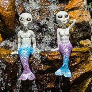 May include: Two gray alien mermaid figurines with blue and purple tails, sitting on a rock with a waterfall in the background.