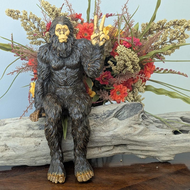 May include: A dark brown, furry Bigfoot figurine with a light-colored face and feet, sitting in a pose with one hand raised in a peace sign gesture. The creature has textured, shaggy fur detailing across its entire body and a neutral facial expression.