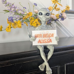 Puede incluir: Una figurita de extraterrestre gris sosteniendo una tarjeta que dice "Feliz cumpleaños Alyssa" con un pastel y velas. La figurita está sentada sobre una superficie negra.