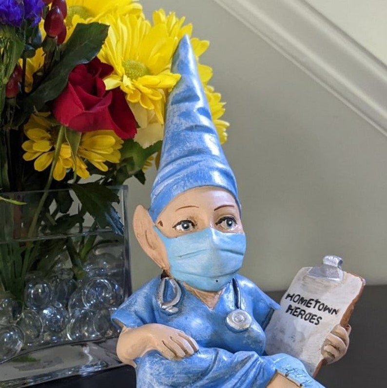 Op de afbeelding: Een blauwe en witte gnoomfiguur die een blauw chirurgisch masker en een blauwe en witte hoed draagt. De gnoom houdt een klembord vast met de tekst "Hometown Heroes" erop. De gnoom zit op een tafel met een vaas met bloemen erachter.