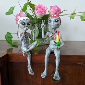 K&ouml;nnte beinhalten: Zwei graue Alien-Figuren mit Friedenssymbolen und einem gr&uuml;n-roten Bong. Das Alien links h&auml;lt eine Zigarette. Das Alien rechts h&auml;lt einen Bong. Die Figuren sitzen auf einer Holzoberfl&auml;che.