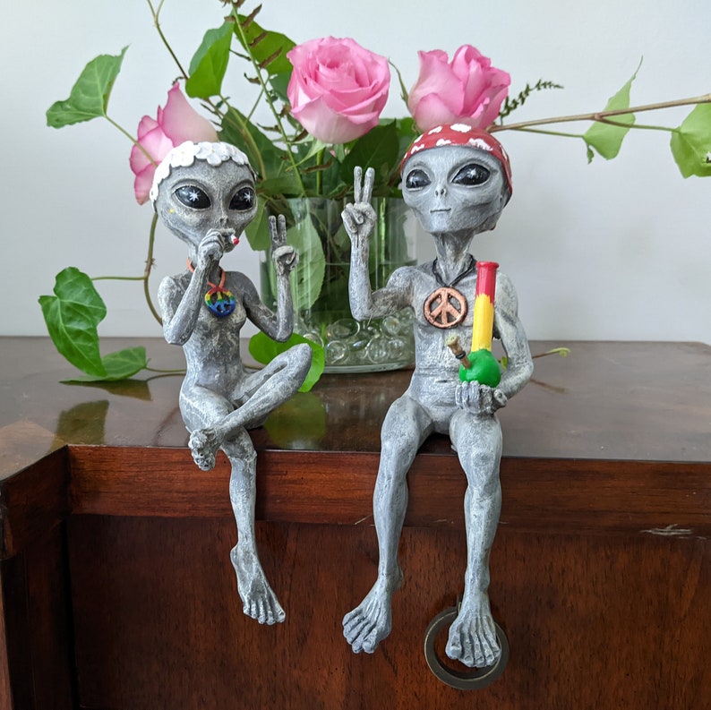 K&ouml;nnte beinhalten: Zwei graue Alien-Figuren mit Friedenssymbolen und einem gr&uuml;n-roten Bong. Das Alien links h&auml;lt eine Zigarette. Das Alien rechts h&auml;lt einen Bong. Die Figuren sitzen auf einer Holzoberfl&auml;che.