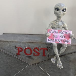May include: A grey alien figure holding a sign saying "Roman Michael Be My Valentine." The alien is perched on a metal mailbox with the word "POST" in red. The sign has a pink background with heart shapes.