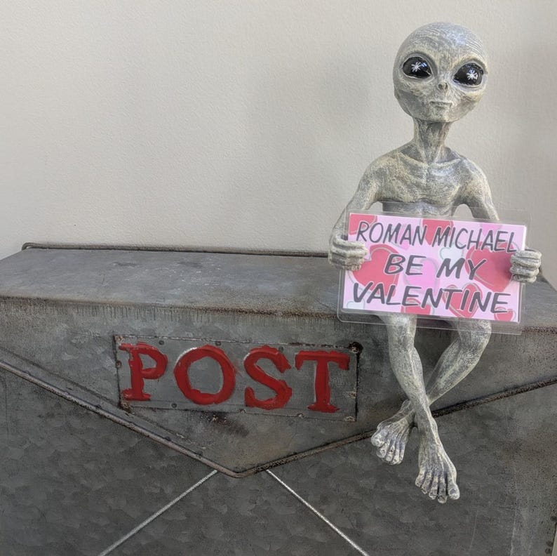 May include: A grey alien figure holding a sign saying "Roman Michael Be My Valentine." The alien is perched on a metal mailbox with the word "POST" in red. The sign has a pink background with heart shapes.