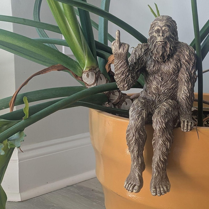 May include: A brown, furry Bigfoot figurine sitting on the edge of a terracotta pot, giving the middle finger.