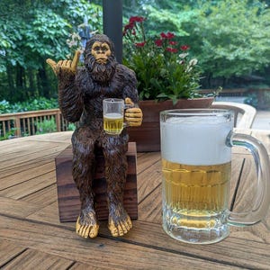 May include: A brown Bigfoot statue with gold accents, holding a small glass of beer, sits on a wooden block. A large glass beer mug filled with beer and foam is next to the statue. The statue is giving a hand gesture.