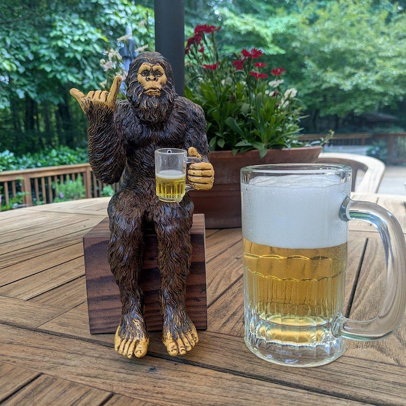 May include: A brown Bigfoot statue with gold accents, holding a small glass of beer, sits on a wooden block. A large glass beer mug filled with beer and foam is next to the statue. The statue is giving a hand gesture.