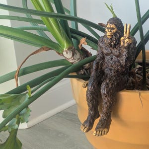 May include: A brown, textured figurine of a hairy creature resembling a Bigfoot or Sasquatch, depicted in a seated position with its legs dangling over the edge. The creature has a detailed face and is making a peace sign gesture with one hand.