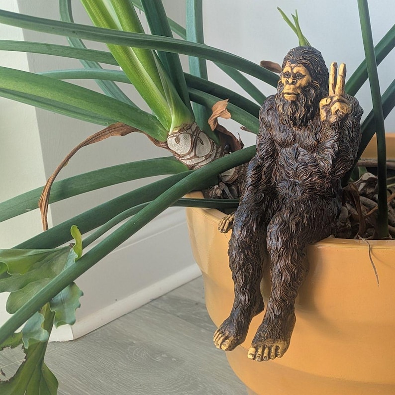 May include: A brown, textured figurine of a hairy creature resembling a Bigfoot or Sasquatch, depicted in a seated position with its legs dangling over the edge. The creature has a detailed face and is making a peace sign gesture with one hand.