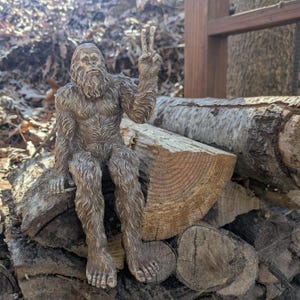 May include: A brown, textured figurine of a hairy, bipedal creature resembling a Bigfoot or Sasquatch. The figure is depicted in a seated position with one arm raised, displaying a peace sign gesture with its fingers, and has a detailed fur texture.