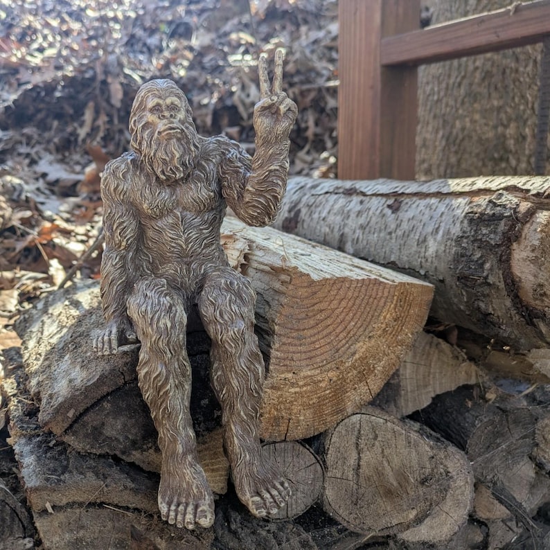 May include: A brown, textured figurine of a hairy, bipedal creature resembling a Bigfoot or Sasquatch. The figure is depicted in a seated position with one arm raised, displaying a peace sign gesture with its fingers, and has a detailed fur texture.