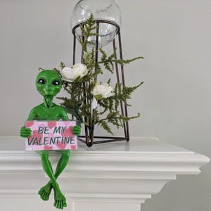 May include: A green alien ornament holding a sign saying "BE MY VALENTINE" with pink hearts. The alien sits on a white surface, with a glass globe and floral display in the background.